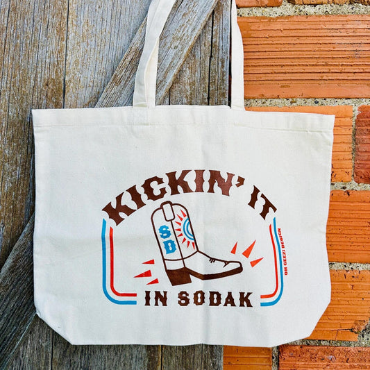 Tote bag with 'Kickin' It in Sodak' design on a wooden and brick background