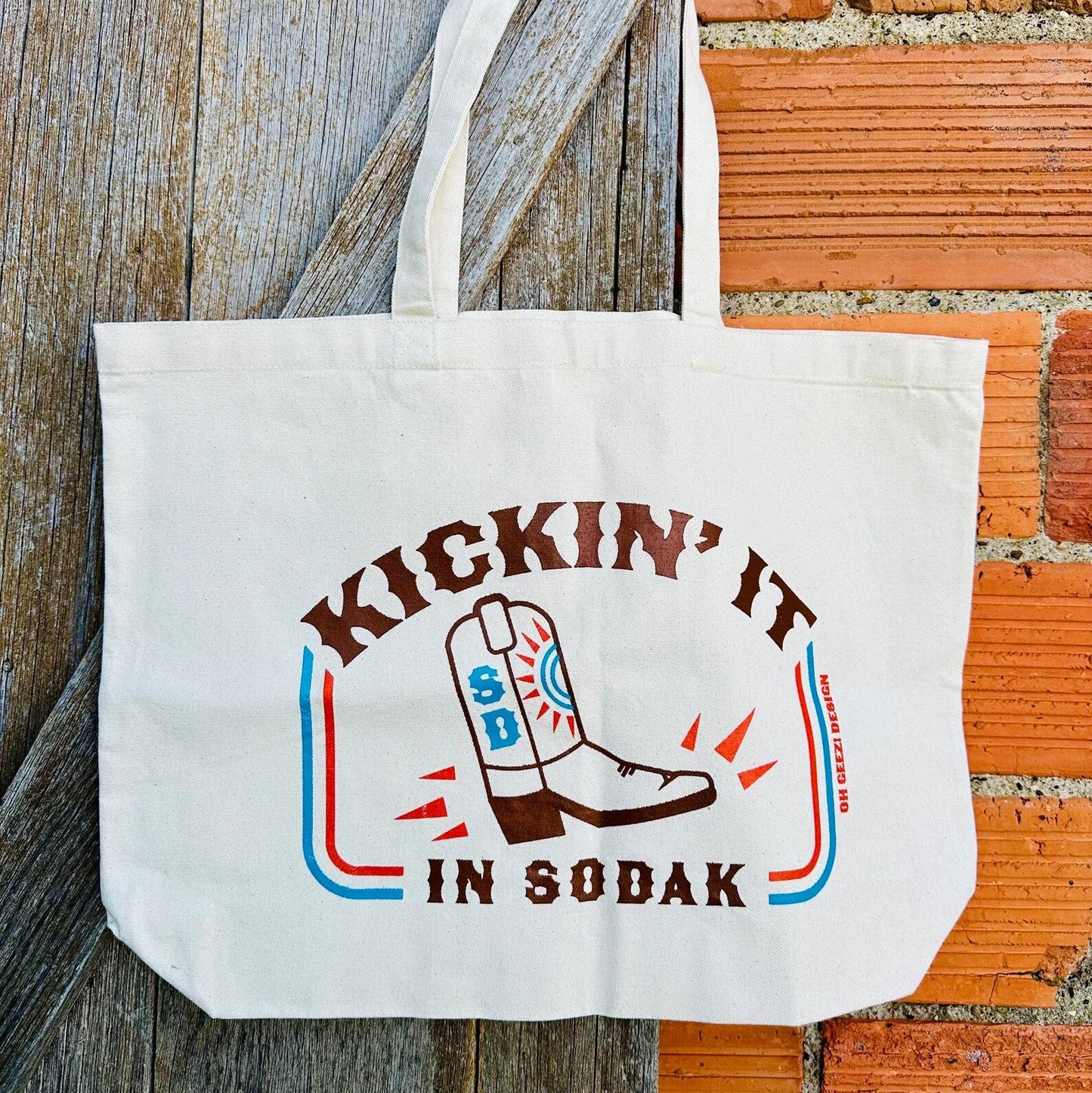Tote bag with 'Kickin' It in Sodak' design on a wooden and brick background