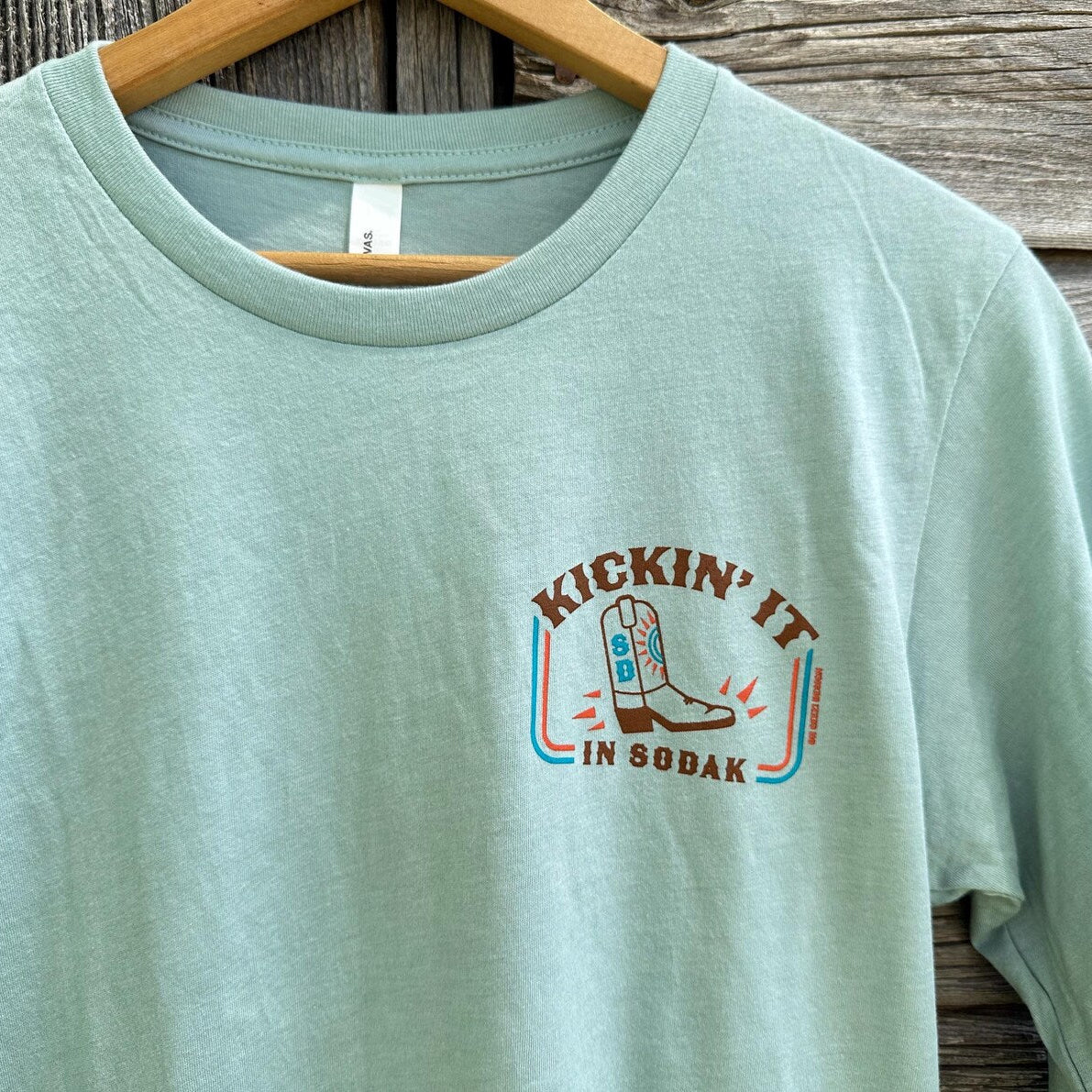 Light blue t-shirt with 'Kickin' It in Sodak' graphic on a wooden background