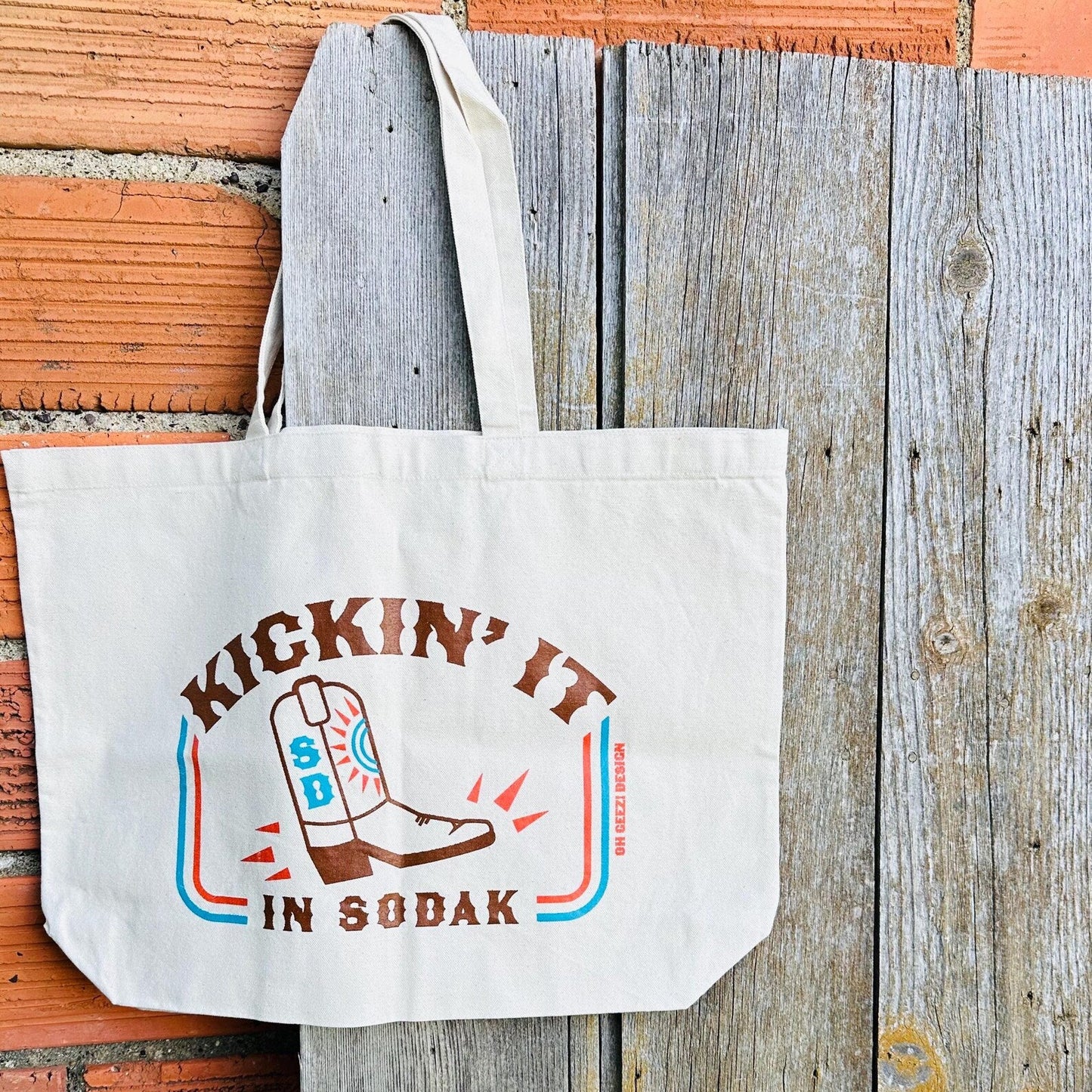 Canvas tote bag with 'Kickin' it in So Dak' design on a wooden surface
