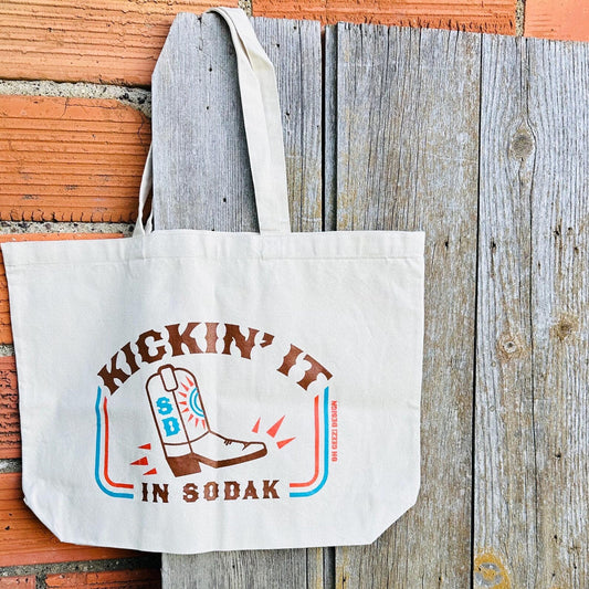 Canvas tote bag with 'Kickin' it in So Dak' design on a wooden surface