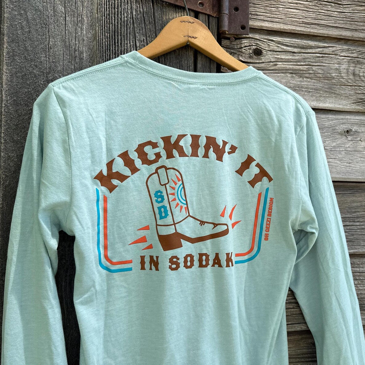 Light blue long-sleeve shirt with a graphic design of a boot and text 'Kicking It in Sodak' on a wooden background.