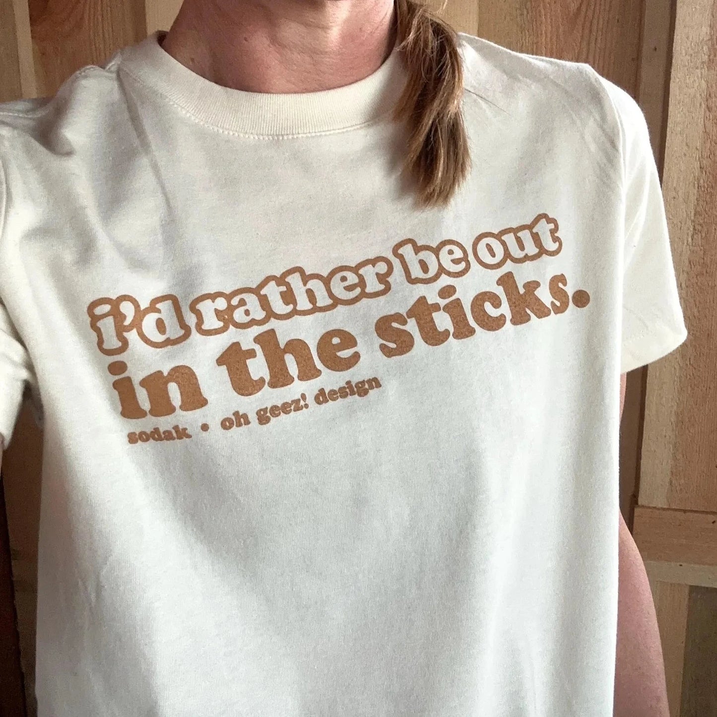 Person wearing a white t-shirt with text 'I'd rather be out in the sticks' in front of a wooden background.