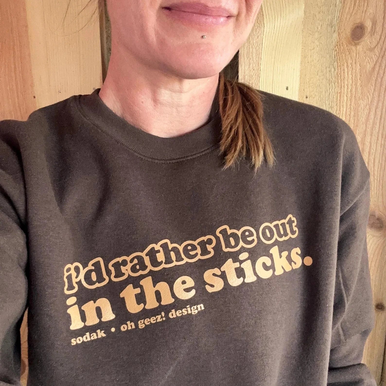 Person wearing a brown sweatshirt with text 'I'd rather be out in the sticks' in front of a wooden wall.