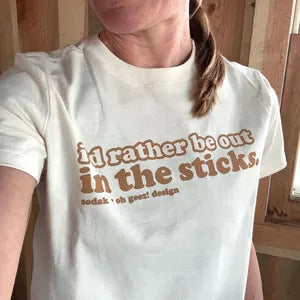 Person wearing a white t-shirt with text 'I'd rather be out in the sticks' in a wooden interior setting.